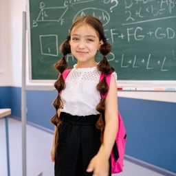 Young girl with a pink backpack standing in front of a chalkboard at school.